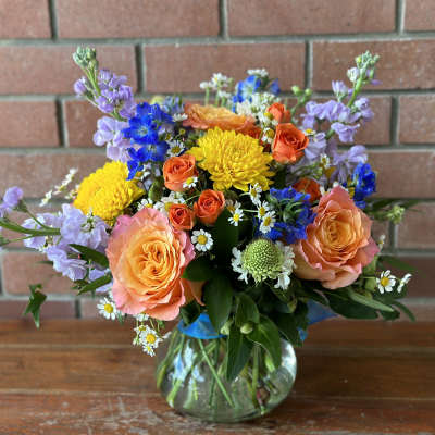 Mixed bouquet of orange roses, yellow mums, blue flowers and white daisies in a round glass vase