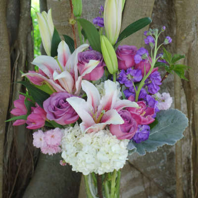 Tall glass vase of pink lilies, purple roses, white hydrangeas, and mixed pink and purple flowers.