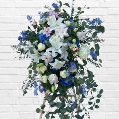 Tall floral spray with white lilies, blue blooms, and trailing greenery on an easel