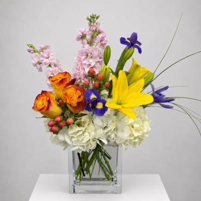 Mixed bouquet in a clear square vase with yellow lilies, orange roses, and blue irises