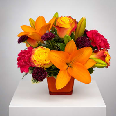 Orange lilies, yellow roses, and pink carnations in an amber vase