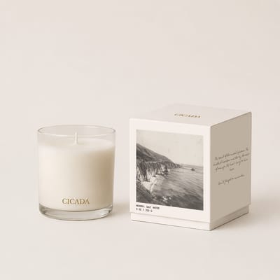 White candle in a glass tumbler beside a boxed candle