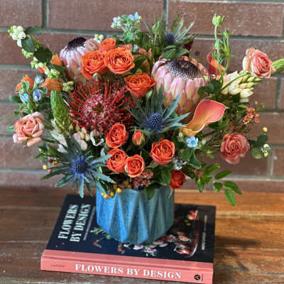 Colorful arrangement with orange roses, protea, and calla lilies in a blue vase on books