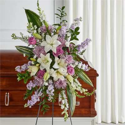 Standing spray of white lilies, lavender blooms, and pink roses displayed before a wooden casket.