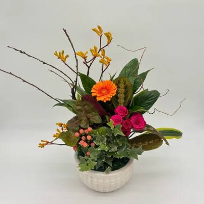 Asymmetrical arrangement with orange gerbera, hot pink roses, berries and foliage in a white ceramic pot