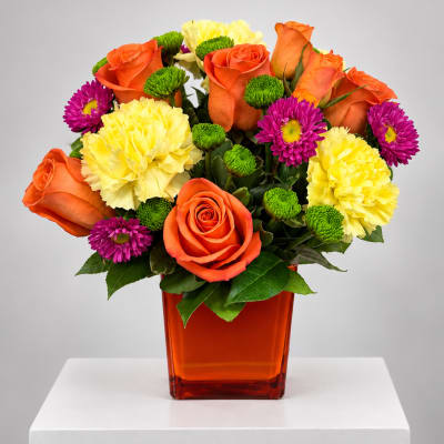 Orange roses, yellow carnations, and purple daisies in an orange vase