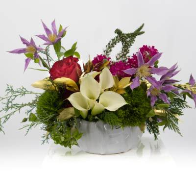 Low arrangement of white calla lilies, red roses, and purple flowers in a white ceramic bowl