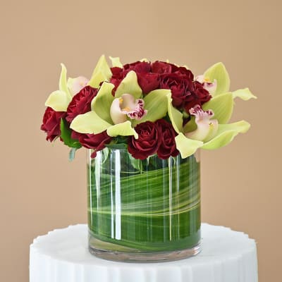 Red roses and pale green orchids arranged in a glass cylinder vase.