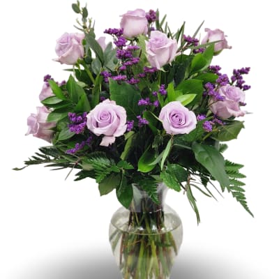 Lavender roses and small purple flowers arranged with lush foliage in a clear glass vase