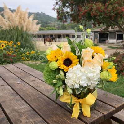 Bouquet of sunflowers, white hydrangea, and pale roses in a glass vase with a yellow ribbon.