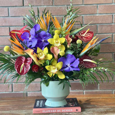 Tall tropical arrangement of orange, purple, yellow, and red flowers in a pale green vase