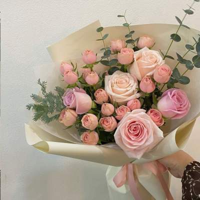 Hand-tied bouquet of light pink and peach roses and rosebuds wrapped in cream paper with a pink ribbon