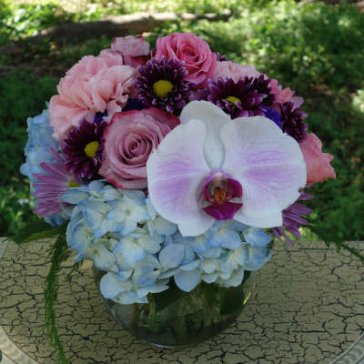 Round arrangement of pink roses and carnations with blue hydrangeas and a white orchid in a clear glass bowl
