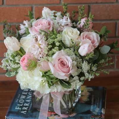 Low pink and white rose arrangement in a clear glass cube vase with ribbon, set on books.