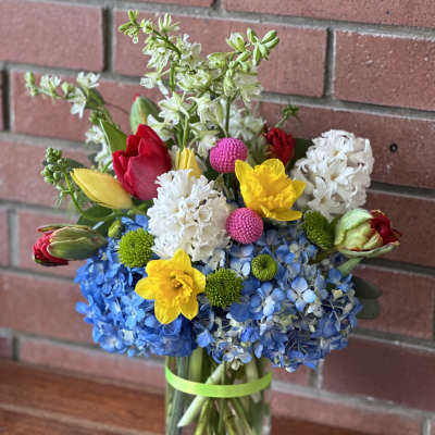 Bright mixed bouquet with blue hydrangeas, yellow daffodils, tulips, and white flowers in a clear glass vase