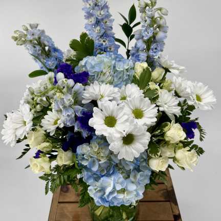 White daisies, pale blue hydrangeas, and cream roses arranged with blue delphinium in a clear glass vase.