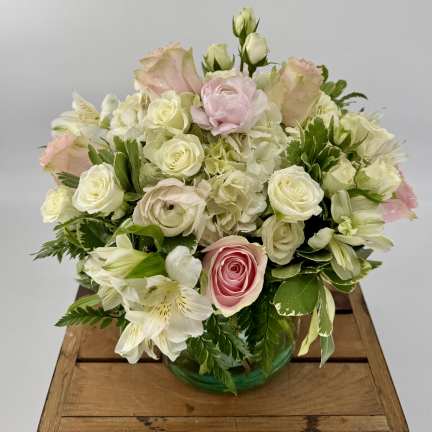 Compact arrangement of white and pale pink roses, hydrangeas, and lilies in a round glass vase