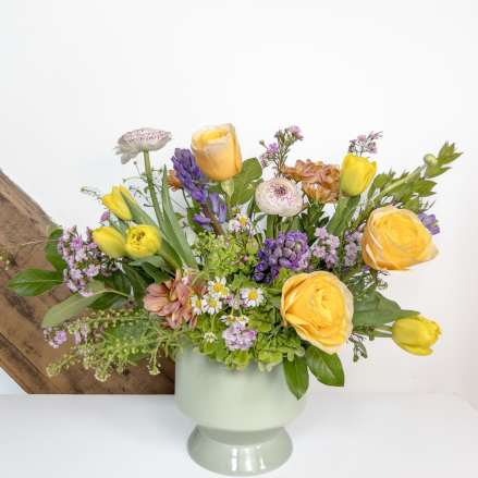 Low arrangement of yellow roses, tulips, and mixed pastel flowers in a pale green pedestal vase