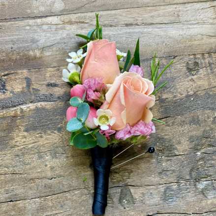 Peach rose boutonniere with pink and white accent flowers wrapped in black ribbon on rustic wood.
