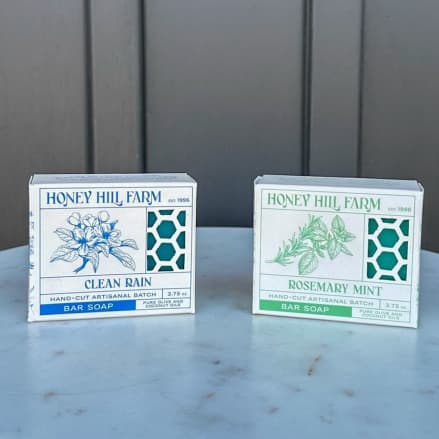 Two Honey Hill Farm bar soap boxes, Clean Rain and Rosemary Mint, on a light stone surface