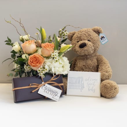 Peach and white flower arrangement with teddy bear, chocolates, and a baby welcome card.