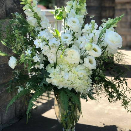 Tall all-white floral arrangement with roses, hydrangeas, and snapdragons in a clear glass pedestal vase