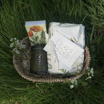 Woven basket gift set with green candle, floral print towel, card, and small white blossoms in grass.