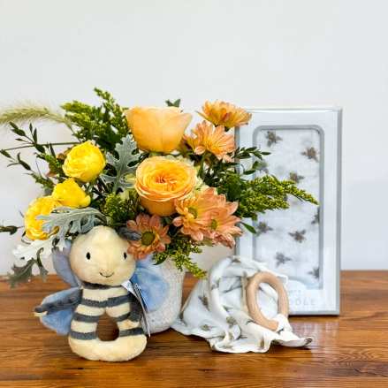 Baby-themed gift set with yellow and peach flowers in a vase, bee plush toy, and bee-print blanket.