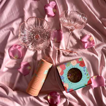 Pink glass coupe and bowl with packaged flower petals on pink fabric with scattered rose petals
