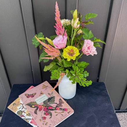 Pink and yellow flowers arranged in a white vase beside floral notebooks.