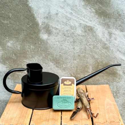 Gardener gift set with black watering can, bar soap, tin, and hand tools on a wooden surface