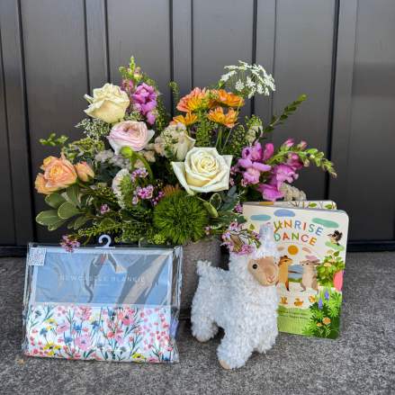Floral arrangement with pastel roses and mixed blooms beside a baby blanket, board book, and plush llama toy
