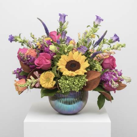 Mixed bouquet with a sunflower, pink roses, and purple blooms in a ribbed iridescent vase