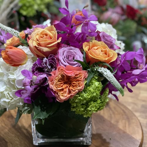 Bright mixed arrangement of orange roses, purple orchids, and hydrangeas in a glass cube vase