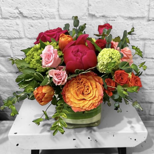 Low arrangement of orange and pink roses, peonies, and tulips in a square glass vase