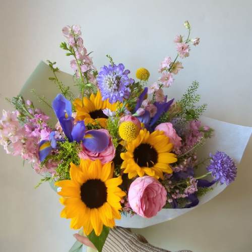 Handheld bouquet with sunflowers, pink roses, and purple blooms wrapped in paper