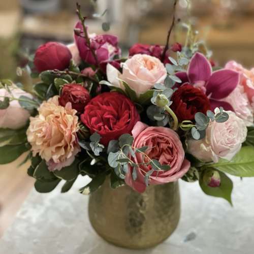 Low gold vase with pink, red, and peach roses and orchids arranged in a lush rounded bouquet.