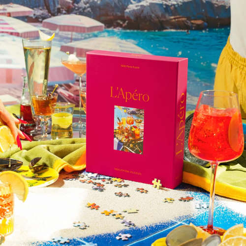 Bright pink L’Apéro jigsaw puzzle box on a sand-like surface with cocktails and puzzle pieces around it.