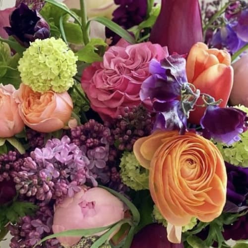 Vibrant mixed arrangement with pink roses, orange ranunculus, tulips, and purple blooms.