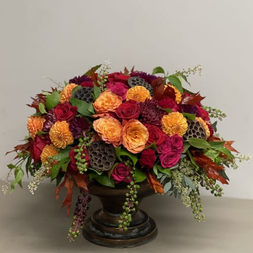 Low arrangement of orange and red roses with dark accent flowers in a bronze pedestal bowl