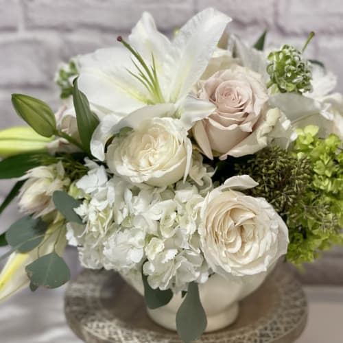 Low white bowl arrangement with white lilies, cream roses, hydrangeas, and green accents