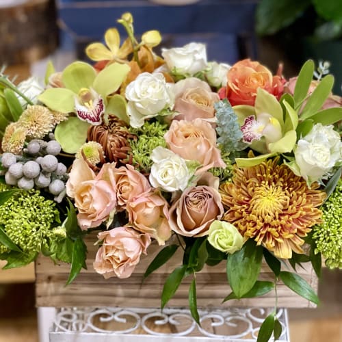 Low wooden box arrangement of peach roses, orchids, and mums in warm mixed colors