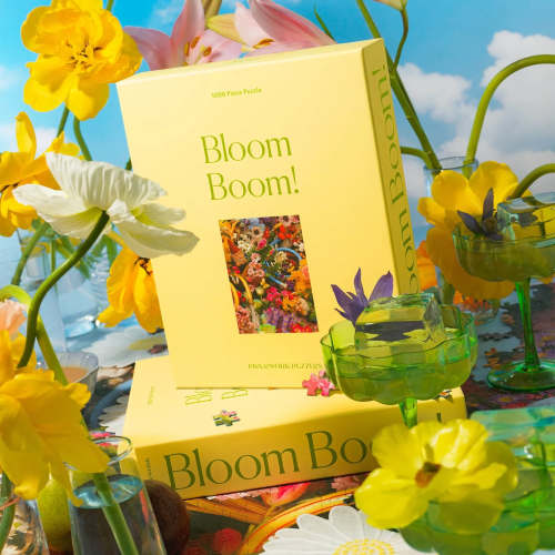Bloom Boom jigsaw puzzle box styled among bright yellow and pink flowers and green glassware.