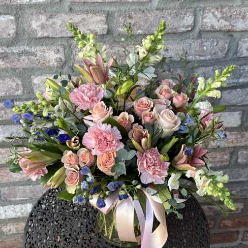 Mixed arrangement of pink roses, carnations, lilies, and blue flowers in a clear vase with a pink ribbon.