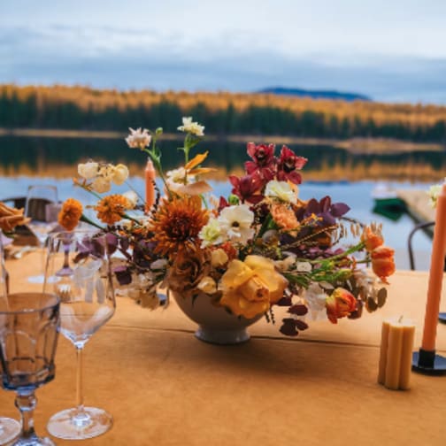 Low centerpiece of yellow and orange flowers in a white bowl on a table by a lake in autumn.