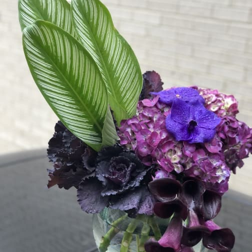 Purple hydrangea and dark calla lilies in a glass vase with striped green leaves
