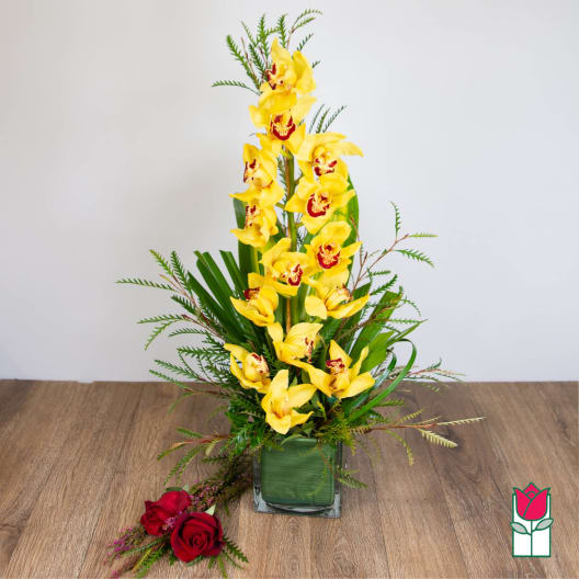 Tall arrangement of yellow orchids in a square glass vase with two red roses laid beside it