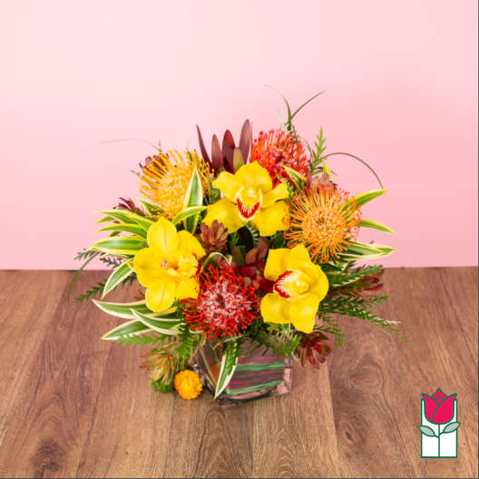 Tropical arrangement of yellow orchids and orange pincushion blooms in a square glass vase.