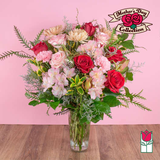 Tall mixed bouquet of red roses and pink flowers in a clear glass vase against a pink background