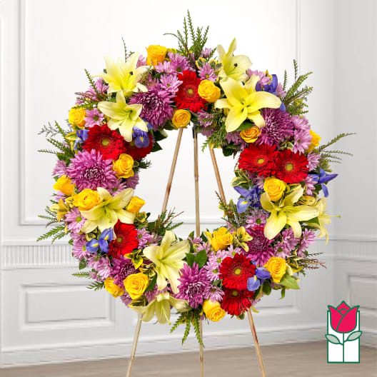 Large floral wreath on a stand with yellow lilies, red gerberas, and purple blooms